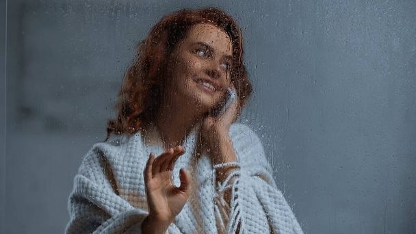 positive and curly woman talking on smartphone behind window glass with raindrops - Фото, изображение