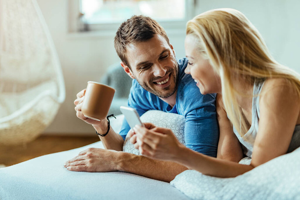 Happy man drinken van koffie in de ochtend en communiceren met zijn vriendin die gebruik maakt van mobiele telefoon in de slaapkamer. - Foto, afbeelding