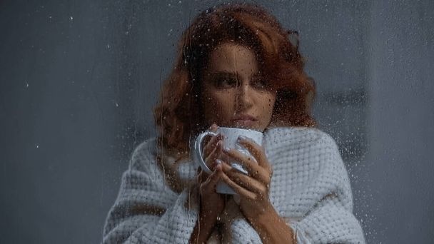 sick woman holding cup with hot beverage behind window glass with raindrops - Foto, Imagem