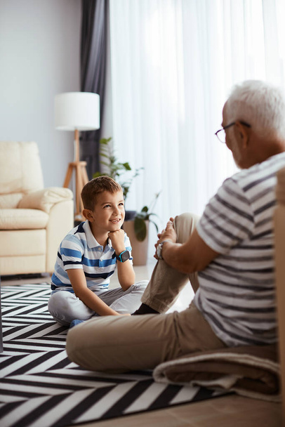 Щасливий маленький хлопчик слухає свого діда, який розмовляє з ним, відпочиваючи на підлозі вдома
.  - Фото, зображення