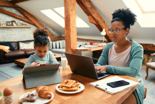 Small black girl eating breakfast and using digital tablet while her mother is working on laptop at dining table.  - Photo, Image