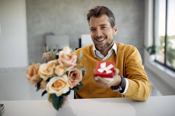Happy man holding a present an bouquet of roses while sitting at office desk and looking at camera. - Photo, Image