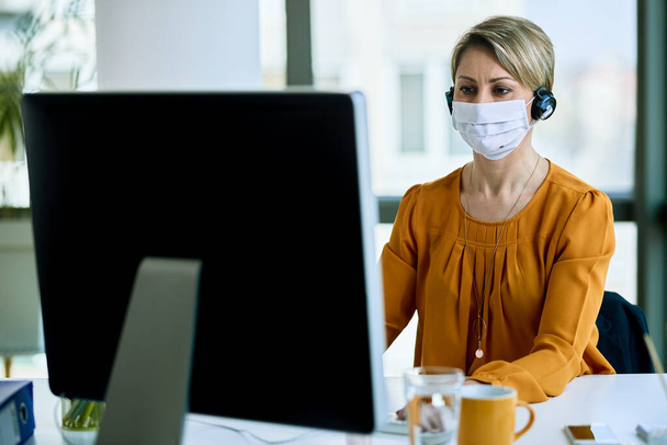 Female entrepreneur wearing headset and protective face mask while working on a computer in the office.  - Foto, imagen