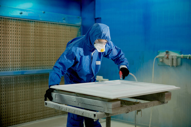 Людина в захисному робочому одязі розпилює фарбу на шматок меблів на деревообробному виробництві
.  - Фото, зображення