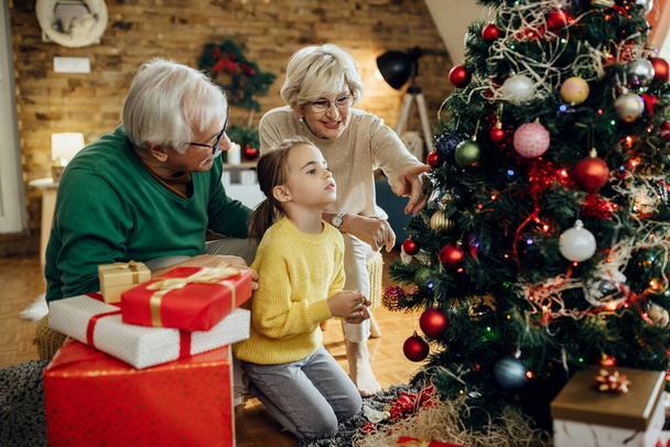 Happy mature couple with granddaughter decorating Christmas tree in the living room.  - Foto, Imagen