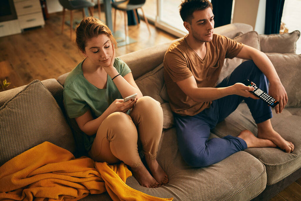 Young couple relaxing on the sofa in the living room. Focus is on woman using mobile phone and feeling bored while her boyfriend is changing channels on a TV. - Foto, immagini