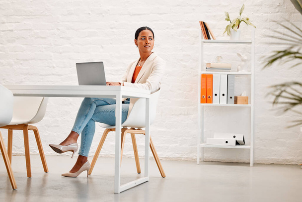 Young mixed race businesswoman thinking while working on a laptop at a table in an office at work. One hispanic businessperson typing on a laptop while sitting in a chair at a desk. - Foto, Bild