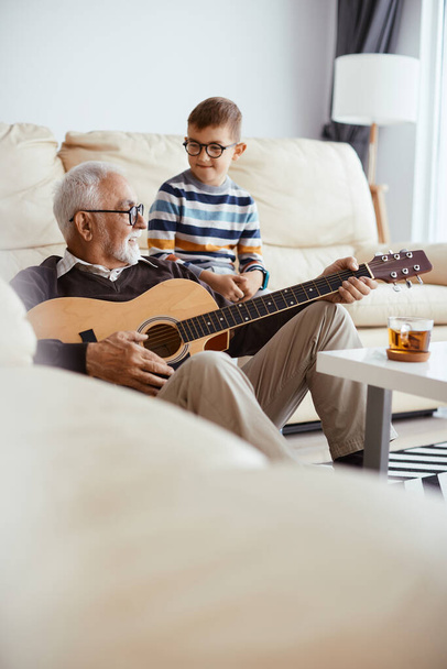 Happy grandfather playing acoustic guitar while spending time with his grandson at home.  - Foto, afbeelding