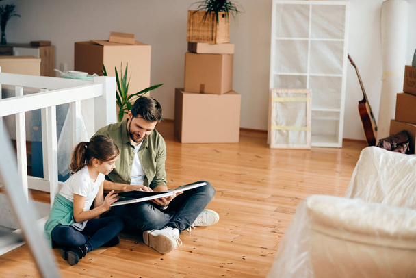 Улыбающиеся отец и дочь читают книгу, сидя на полу в своем новом доме.  - Фото, изображение