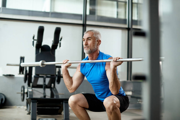 Mature sportsman exercises strength while lifting barbell in gym - Foto, imagen