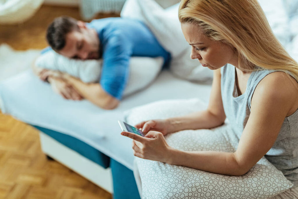 Young worried woman using smart phone while her boyfriend is napping on the bed.  - Valokuva, kuva