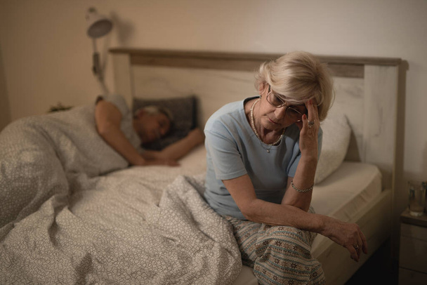 Worried mature woman thinking of something while sitting in bedroom at night. Her husband is sleeping in the background.  - Foto, Bild