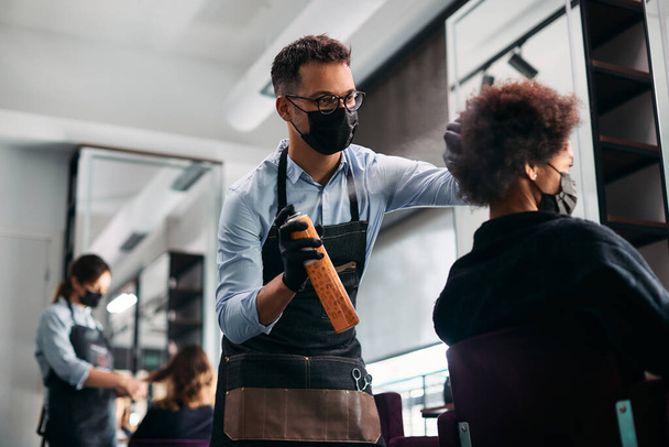 Male hairdresser styling African American woman's hair with hairspray and wearing face mask due to coronavirus pandemic.   - Valokuva, kuva