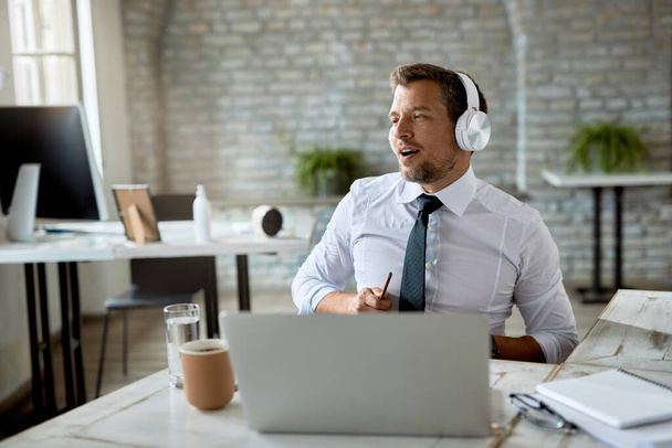 Середній дорослий бізнесмен підписується під час прослуховування музики над навушниками і роботи на ноутбуці в офісі
.  - Фото, зображення