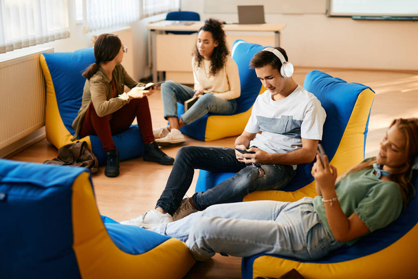 High school student listening music over headphones and using smart phone while relaxing with his classmates in the classroom.  - Photo, Image