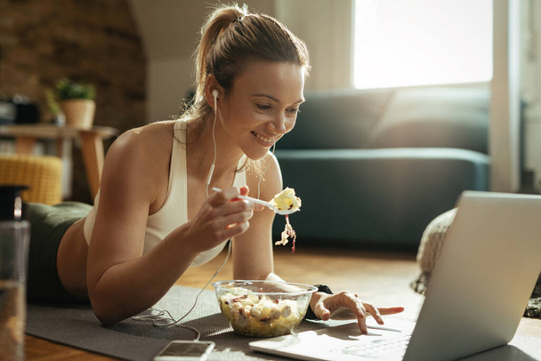 Šťastná atletická žena těší ve zdravém jídle a pomocí notebooku při relaxaci na podlaze.  - Fotografie, Obrázek
