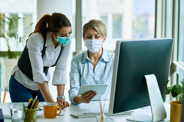 Female entrepreneurs with face masks cooperating while working on touchpad in the office. - Foto, imagen