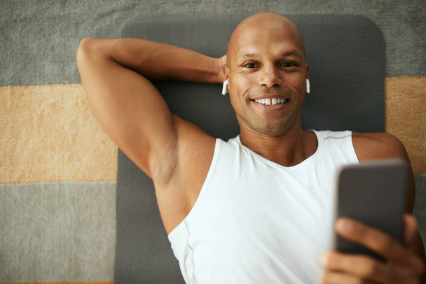 Happy African American athletic man listening music over earphones and using mobile phone while lying down on floor after home workout.  - Fotó, kép