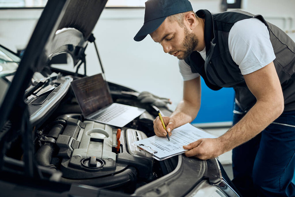 Young mechanic filling paperwork while examining car engine at auto repair shop. - Φωτογραφία, εικόνα