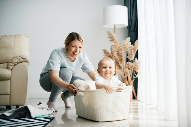 Playful mother pushing her baby boy in white basket while playing with him at home.  - Foto, afbeelding