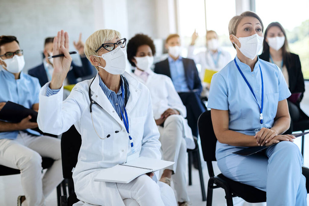 Mature doctor raising hand to ask a question while attending healthcare seminar with group of colleagues and business people at convention center during coronavirus epidemic.  - 写真・画像