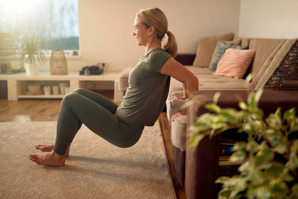 Happy sportswoman leaning on the sofa and practicing triceps dips during home workout. - Fotografie, Obrázek