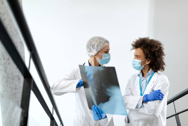 Жінки-радіологи обговорюють під час аналізу рентгенівського зображення пацієнта в лікарняному коридорі. Вони носять захисні маски для обличчя через пандемію коронавірусу
.  - Фото, зображення