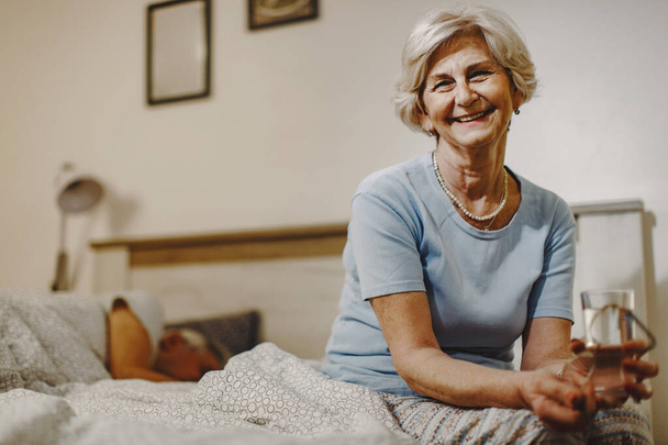 Happy senior woman with glass of water sitting on the bed while her husband is sleeping in the background.  - Fotografie, Obrázek