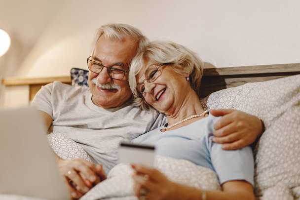 Happy senior couple using laptop and credit card while relaxing in bedroom. - Foto, afbeelding