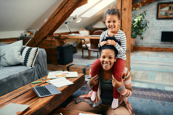 Happy African American working mother having fun with her daughter  at home and looking at camera. Little girl is sitting on mother's shoulders. - Fotoğraf, Görsel