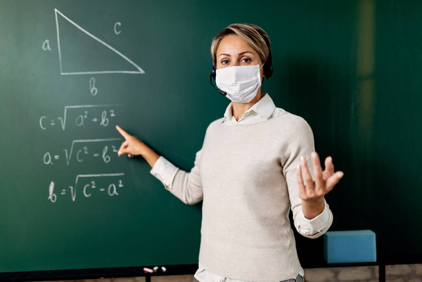 Female teacher with a face mask standing in front of chalkboard and giving online math lecture during coronavirus epidemic. - Photo, image