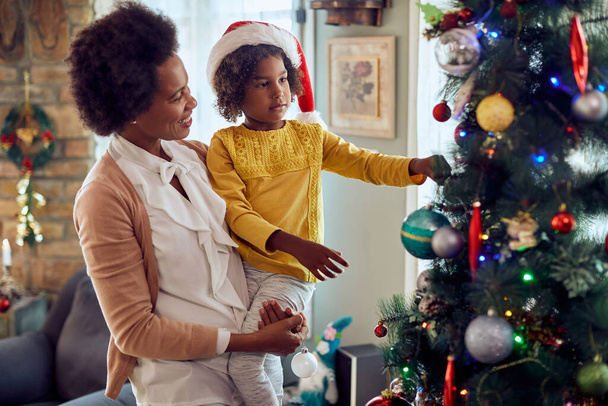 Happy African American mother enjoying while decorating Christmas tree with her small daughter at home.  - Foto, immagini