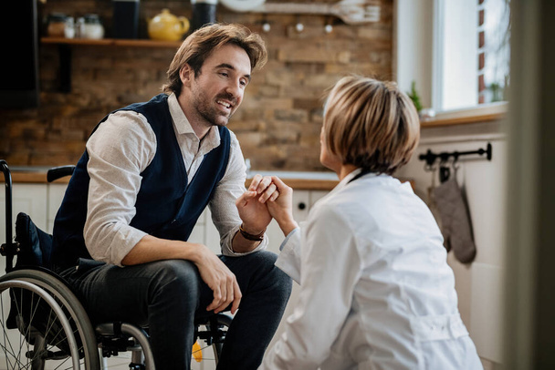 Młody szczęśliwy mężczyzna na wózku inwalidzkim trzymający się za ręce ze swoim lekarzem podczas rozmowy z nią podczas wizyty w domu.  - Zdjęcie, obraz