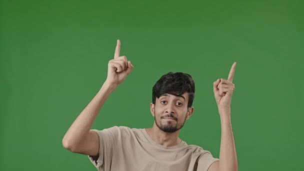 Positive arab man points forefingers up at copy space for advertisement blank place recommends promotional imaginary product pretends shooting pistols choosing make choice isolated on green background - Metraje, vídeo