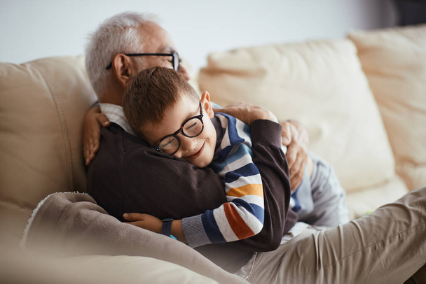 Щасливий маленький хлопчик обіймає свого діда, який розслабляється на дивані вдома
. - Фото, зображення