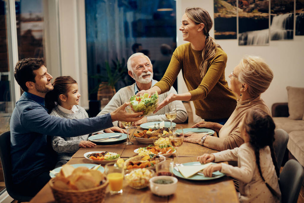 Happy multi-generation family enjoying in a lunch together at home. Focus is on young woman serving salad at dining table.  - Φωτογραφία, εικόνα
