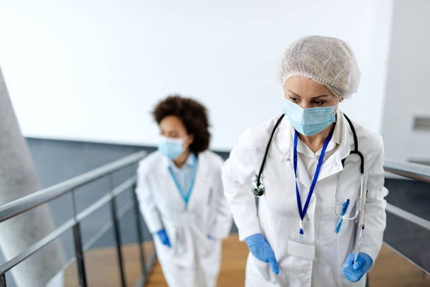 Bezorgde vrouwelijke arts draagt een beschermend gezichtsmasker en loopt naar boven in een hal van de medische kliniek. Haar collega is op de achtergrond.  - Foto, afbeelding