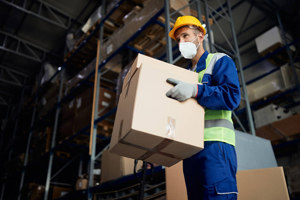 Low angle view of warehouse worker carrying cardboard boxes and wearing face mask due to COVID-19 pandemic. - Photo, image