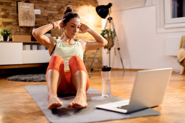 Dedicated sportswoman practicing sit-ups while using laptop and having exercises class at home.  - 写真・画像