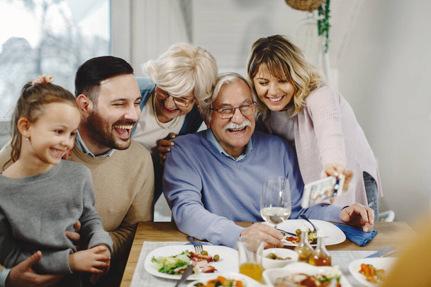 Щаслива розширена сім'я розважається, приймаючи селфі зі смартфоном під час обіду в їдальні
.  - Фото, зображення