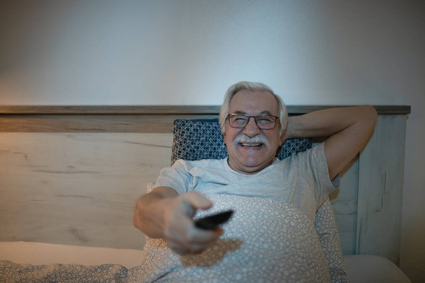 Vrolijke senior man kijkt tv in de slaapkamer 's nachts. - Foto, afbeelding