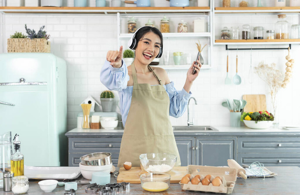 heureux gai jeune asiatique femme élever smartphone poing bonne humeur porter casque écouter de la musique dans la cuisine à la maison. - Photo, image