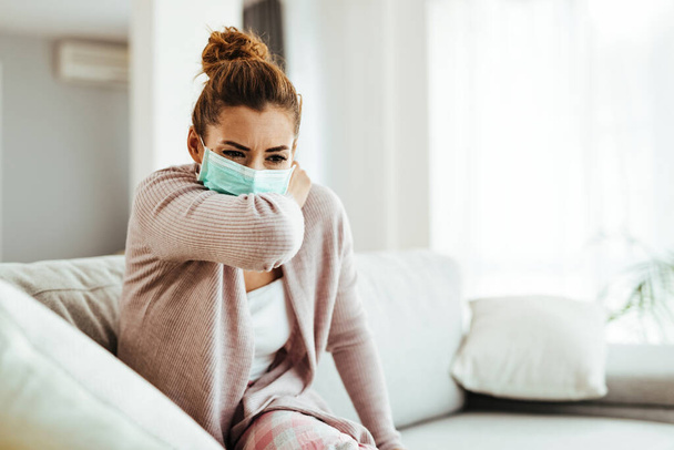 Woman wearing sneezing into her elbow while wearing protective face mask at home.  - Zdjęcie, obraz