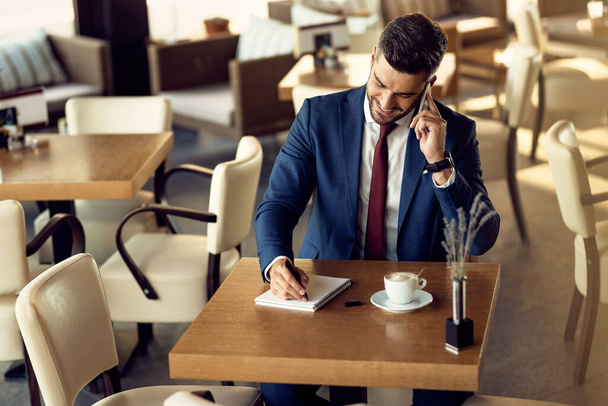  Happy businessman talking on cell phone and writing in notebook while sitting in a cafe.  - Photo, Image