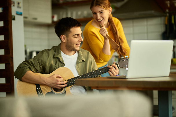 Happy man having fun and playing acoustic guitar while his wife is singing at home. - Φωτογραφία, εικόνα