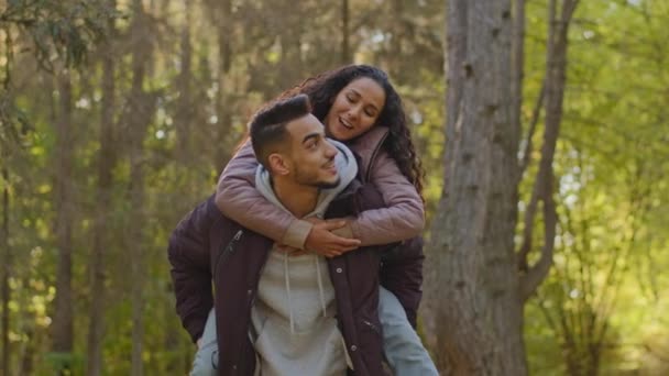 Happy young carefree married couple in love on romantic date in park in nature joyful girl sits on piggyback hugging man by shoulders enjoys freedom with arms outstretched to side talking laughing - Séquence, vidéo