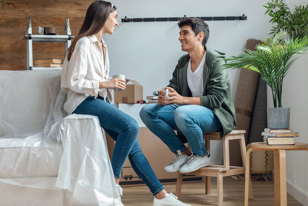 Shot of young beautiful couple drinking a cup of coffe while relaxing from moving in their new house arround cardboard boxes - Zdjęcie, obraz