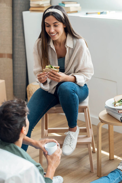 Shot of cute couple eating a sandwich while relaxing from moving in their new house arround cardboard boxes - Zdjęcie, obraz