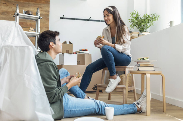 Shot of cute couple eating a sandwich while relaxing from moving in their new house arround cardboard boxes - Photo, image