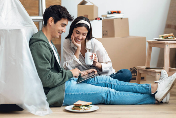 Shot of cute couple using tablet while relaxing from moving in their new house arround cardboard boxes - Foto, imagen